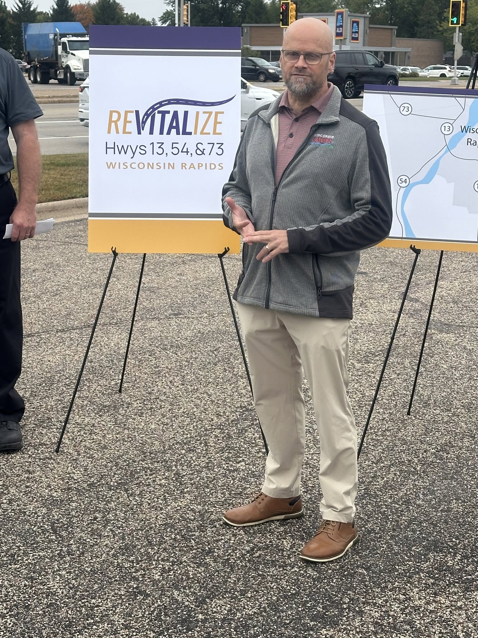 A speaker at a Revitalize Community Highways event in Wisconsin Rapids, beside signage for the Highways 13, 54, and 73 project